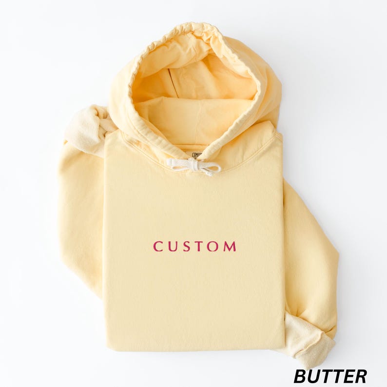 May include: A light yellow hoodie with a drawstring hood and long sleeves. The word "CUSTOM" is printed in burgundy on the front. The word "BUTTER" is printed in black at the bottom right corner. The hoodie is made of a soft material.