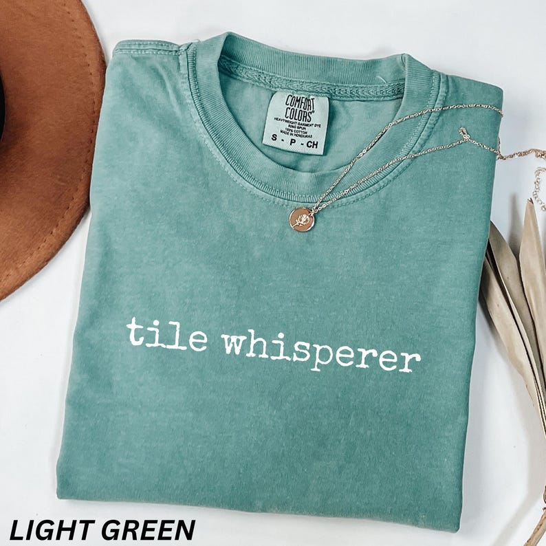 K&ouml;nnte beinhalten: Ein hellgr&uuml;nes T-Shirt mit dem wei&szlig;en Aufdruck "tile whisperer". Eine silberne Halskette mit einem runden Anh&auml;nger liegt auf dem Shirt. Das Shirt ist mit "Comfort Colors" gekennzeichnet und hat die Gr&ouml;&szlig;e Small.