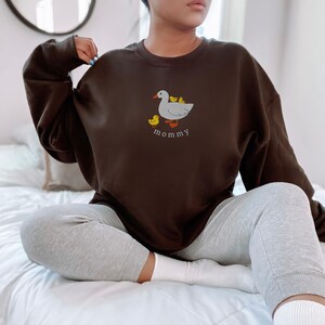 May include: A brown sweatshirt with a white duck and yellow ducklings embroidered on the front. The text "mommy" is embroidered below the duck family.