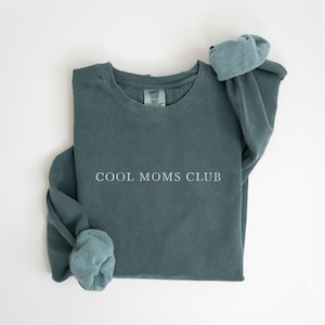 May include: A teal sweatshirt with the white text "COOL MOMS CLUB". The sleeves are turned up, showing a lighter teal lining. The garment is made of a soft material. A casual top.