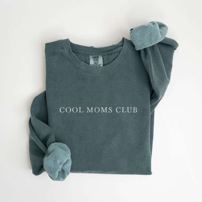 May include: A teal sweatshirt with the white text "COOL MOMS CLUB". The sleeves are turned up, showing a lighter teal lining. The garment is made of a soft material. A casual top.