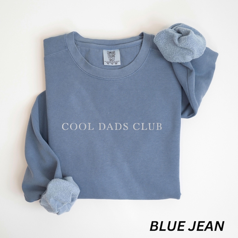 May include: A blue jean-colored sweatshirt with the text "COOL DADS CLUB" in white. The sweatshirt has a crew neck and long sleeves. The sleeves are cuffed. The brand tag is visible at the top. The words "BLUE JEAN" are at the bottom.