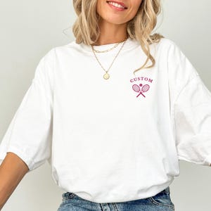 May include: White oversized t-shirt with a small magenta tennis racket and ball graphic. The word "CUSTOM" is above the graphic. The shirt is made of a soft material and has a relaxed fit.