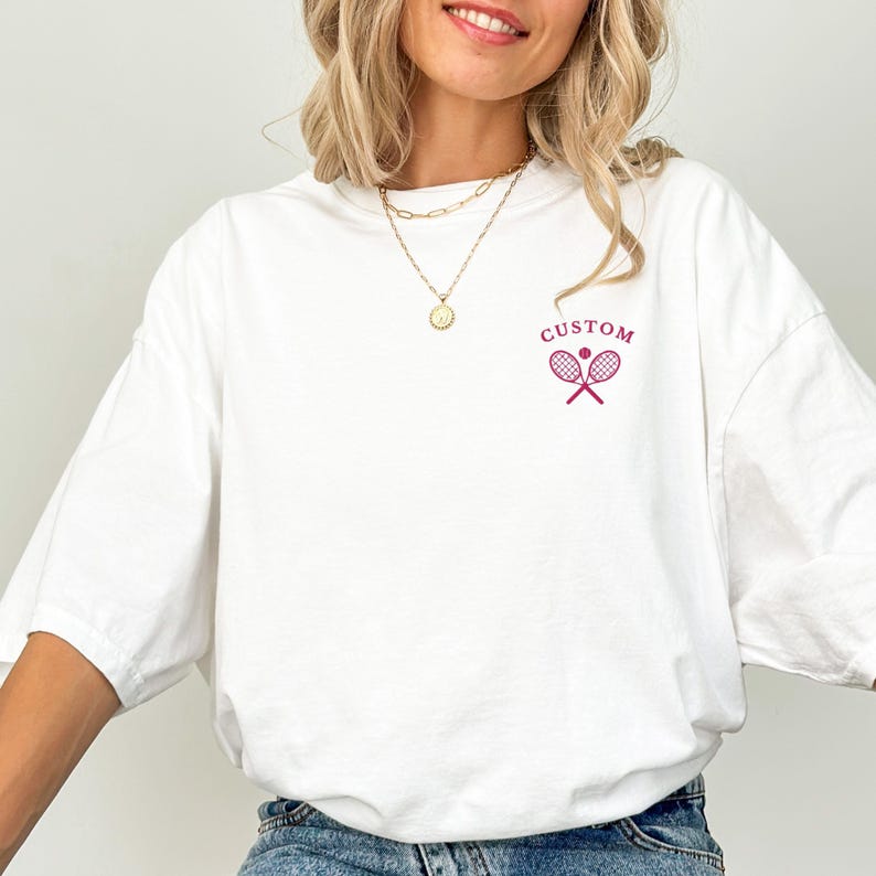 May include: White oversized t-shirt with a small magenta tennis racket and ball graphic. The word "CUSTOM" is above the graphic. The shirt is made of a soft material and has a relaxed fit.