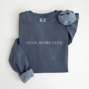 May include: A slate blue sweatshirt with the text "COOL MOMS CLUB" in white. The long sleeves are cuffed, and a matching scrunchie rests on the shoulder. The sweatshirt is made of a soft, comfortable material.