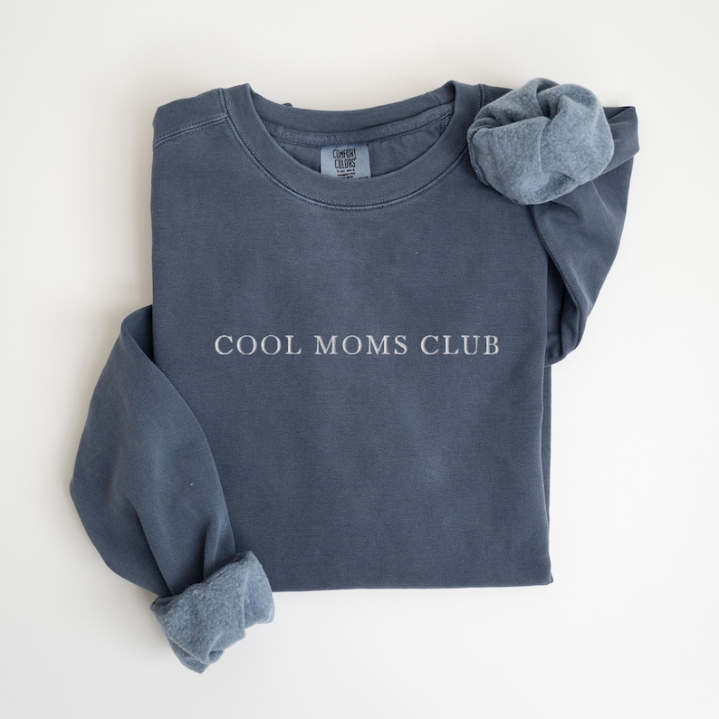 May include: A slate blue sweatshirt with the text "COOL MOMS CLUB" in white. The long sleeves are cuffed, and a matching scrunchie rests on the shoulder. The sweatshirt is made of a soft, comfortable material.