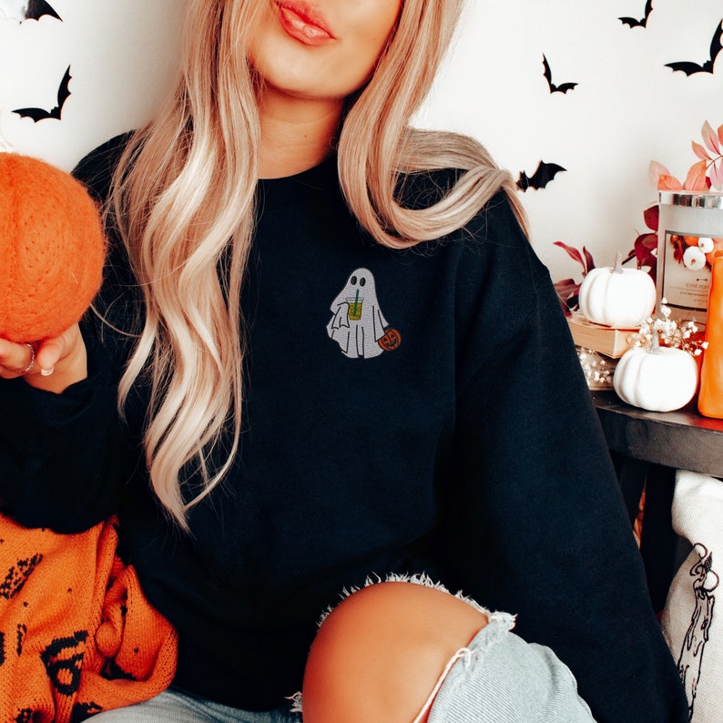 May include: Black sweatshirt with a white ghost holding a green beverage and a small orange pumpkin. The ghost is embroidered on the front of the sweatshirt.
