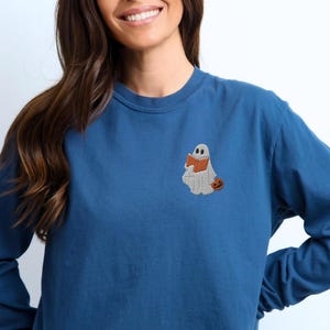 K&ouml;nnte beinhalten: Ein blaues Sweatshirt mit langen &Auml;rmeln und einem gestickten Design eines Geistes, der ein Buch liest und einen K&uuml;rbis h&auml;lt.