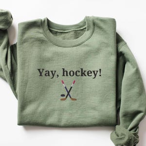 Puede incluir: Una sudadera verde con el texto "Yay, hockey!" y un gr&aacute;fico de dos palos de hockey cruzados y un disco.
