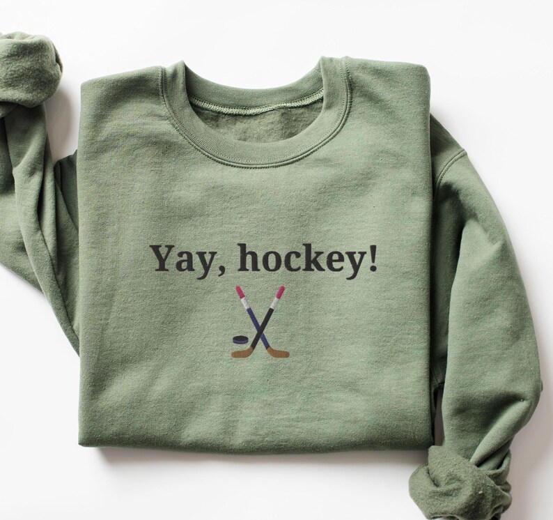 Puede incluir: Una sudadera verde con el texto "Yay, hockey!" y un gr&aacute;fico de dos palos de hockey cruzados y un disco.