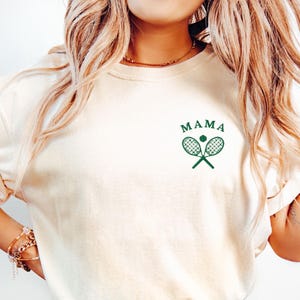 May include: A white t-shirt with a green tennis racket and ball graphic and the text "MAMA" on the front.
