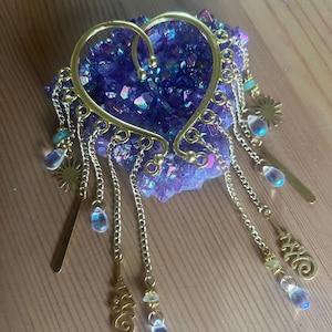 May include: A gold-toned ear cuff shaped like a heart, featuring multiple dangling chains with small sun charms, teardrop-shaped iridescent beads, and decorative metal filigree accents. The ear cuff is resting on a cluster of purple crystals.