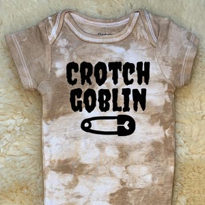 Op de afbeelding: Bruin en witte tie-dye babybody met de zwarte tekst "CROTCH GOBLIN" en een zwarte veiligheidsspeld. De body heeft korte mouwen en drukknoopsluitingen.