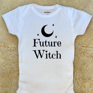 May include: White baby bodysuit with the text "Future Witch" in black script, along with a crescent moon and stars graphic. The bodysuit has short sleeves and snap closures.