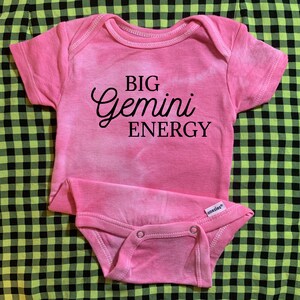 May include: Pink baby onesie with a black tie-dye pattern and the text "BIG Gemini ENERGY" printed on the front.