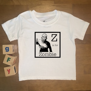 Puede incluir: Camiseta blanca para bebé con una ilustración en blanco y negro de un zombie. La ilustración está en un marco cuadrado con la letra "Z" y el texto "is for Zombie".