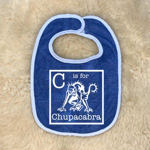 Op de afbeelding: Blauwe baby slabbetje met een lichtblauwe rand. Het slabbetje heeft een witte vierkant met de tekst "C is for Chupacabra" en een cartoon illustratie van het mythische wezen. Het slabbetje is ontworpen voor baby's en peuters.