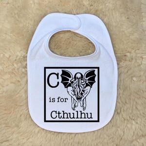 May include: White baby bib with a black graphic of Cthulhu and the text "C is for Cthulhu". The bib has a rounded shape with a snap closure at the neck. The bib is made of a soft material.