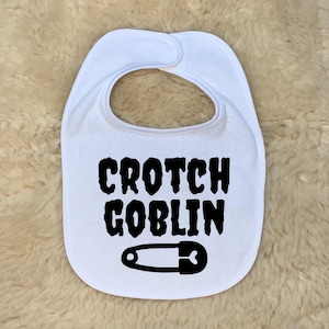 Puede incluir: Babero blanco para bebé con las palabras "Crotch Goblin" en letras negras en negrita. Un gráfico de imperdible negro está debajo del texto. El babero tiene una forma curva con una abertura para el cuello y un cierre a presión.