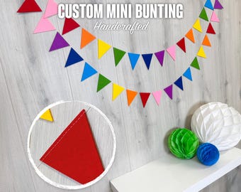 May include: Custom mini bunting in a rainbow of colors, handcrafted. The bunting features small felt triangle flags in red, pink, purple, blue, green, yellow, and orange. The image also shows a close-up of a red flag and decorative paper balls.