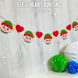 May include: A handmade felt elf and heart bunting with red hearts and green elves. The bunting is strung on a white string and has the text "ELF & HEART BUNTING Handmade" at the top.