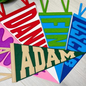Puede incluir: Una colección de banderines de fieltro coloridos, cada uno con un nombre en letras en negrita. Los banderines son de varios colores, incluyendo rojo, azul, verde y rosa, con nombres como "DAN", "FIN", "JOSE" y "ADAM". Cada banderín tiene un nombre de color contrastante.