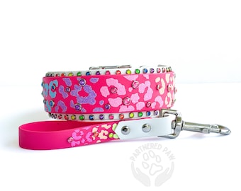 May include: A pink dog collar and leash set with a leopard print design. The collar and leash are adorned with colorful rhinestones. The set includes a white leather accent and a silver metal clasp.