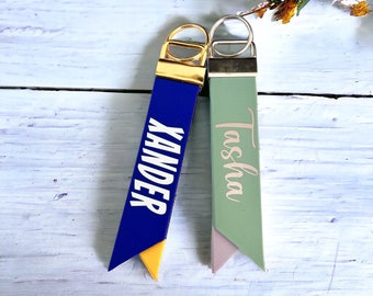 May include: Two personalized keychains. One is blue with the name "XANDER" in white, and the other is green with the name "Tasha" in a cursive font. Both have metal hardware and a contrasting color at the bottom.