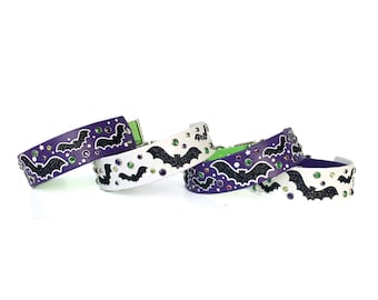 May include: Four purple and white dog collars with black bat silhouettes and colorful rhinestones. The collars are stacked on top of each other.