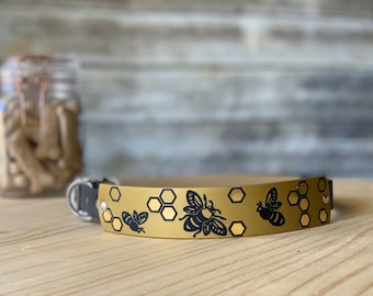 Puede incluir: Un collar de perro de cuero dorado con un diseño de abeja y panal negro.