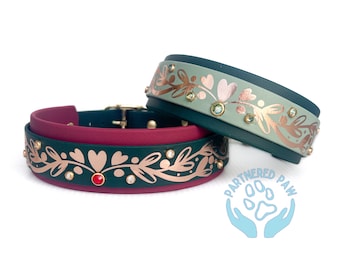 May include: Two dog collars with a green and burgundy base, featuring a gold floral design with rhinestones. The collars are decorated with a red and clear rhinestone. The collars are labeled "Partnered Paw".