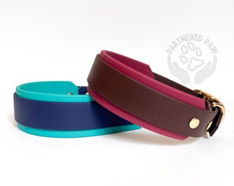 May include: Two pet collars featuring smooth, layered bands. One collar combines a dark blue center with bright turquoise edges. The other collar features a dark brown center with magenta edges, gold-tone hardware, and a Partnered Paw logo.