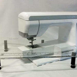 Könnte beinhalten: Eine weiße Nähmaschine mit einem durchsichtigen Lineal aus Kunststoff, das an der Vorderseite befestigt ist. Die Maschine steht auf einer weißen Oberfläche.