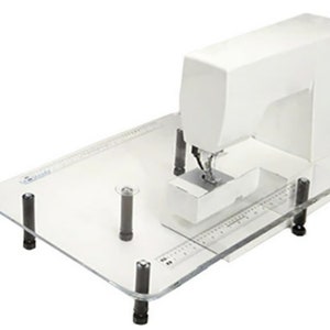Puede incluir: Una máquina de coser blanca sobre una mesa de coser de acrílico transparente con cuatro patas negras. La mesa tiene una regla impresa.