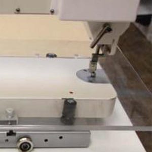 Puede incluir: Primer plano de una máquina de coser blanca con un conjunto de aguja y pie. La máquina está sobre una mesa blanca con una cubierta de plástico transparente.