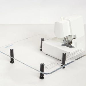 Può includere: Un tavolo per macchina da cucire in acrilico trasparente con quattro gambe regolabili nere. Il tavolo ha un righello stampato sulla superficie e il testo "Sew Steady" stampato nell'angolo in alto a sinistra. Una macchina da cucire bianca è appoggiata sul tavolo.