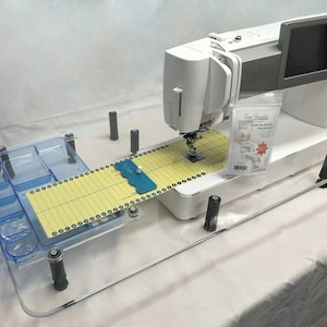 May include: A clear acrylic sewing table extension with a yellow and white ruler, a blue ruler, and a clear plastic storage tray with compartments. The table extension is attached to a white sewing machine with a black and silver screen.