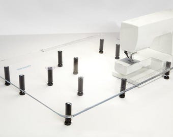 May include: A clear acrylic sewing machine extension table with black adjustable legs. The table is designed to provide extra workspace for sewing projects.