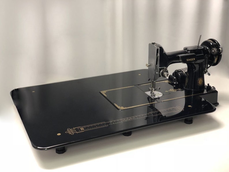 Pode incluir: Uma m&aacute;quina de costura Singer preta com uma base de metal preta e um design dourado. A m&aacute;quina est&aacute; sobre uma mesa preta com uma r&eacute;gua dourada.