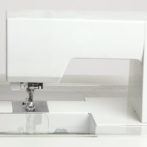 May include: A white sewing machine with a clear acrylic extension table. The machine has a rectangular shape with a needle and presser foot visible. The extension table is 45 cm wide and 30 cm deep.
