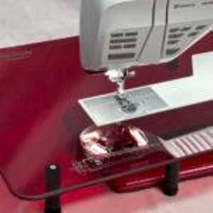 Könnte beinhalten: Ein roter Nähmaschinen-Verlängerungtisch mit einer durchsichtigen Acrylglasplatte. Der Tisch hat einen Metallrahmen und verstellbare Beine. Der Tisch ist so konzipiert, dass er die Nähfläche einer Nähmaschine erweitert.