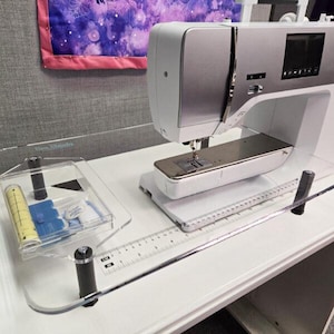 Op de afbeelding: Een witte en zilverkleurige naaimachine op een doorzichtige acryl verlengtafel. De tafel is voorzien van een bedrukte liniaal met centimeter-markeringen. Links staat een doorzichtig accessoirebakje met daarin een geel meetlint.