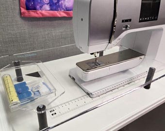May include: A white and silver sewing machine positioned on a clear acrylic extension table. The table features a printed ruler with markings in centimeters and inches. A clear plastic accessory tray sits to the left, containing a yellow measuring tape.