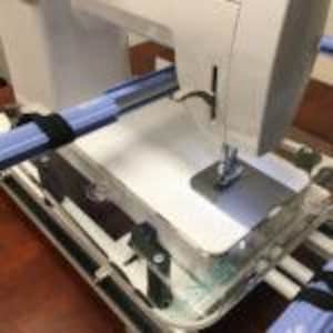 Puede incluir: Una máquina de coser blanca con un aro de bordado azul y negro sujeto al brazo. La máquina está sobre una mesa blanca con una cubierta de plástico transparente.