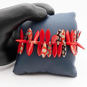 May include: A red and white patterned bracelet with a geometric design. The bracelet is made of small, oval-shaped pieces of ceramic.