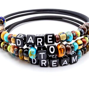 May include: A multi-layered beaded bracelet with black, brown, gold, and blue beads. The bracelet spells out the phrase "Dare to Dream" in black letter tiles.