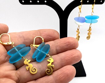 May include: Gold-tone earrings featuring sea glass and seahorse charms. One pair has blue sea glass, the other has blue and white sea glass. The earrings are displayed on a black stand and held in a hand.