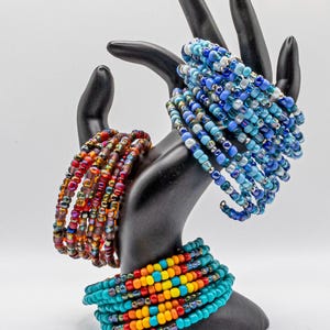 May include: A black hand-shaped display stand with three beaded bracelets. The bracelets are made of colorful glass beads in shades of blue, red, orange, and yellow.