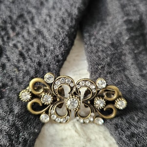 May include: An antique-style brooch with a gold finish. The brooch has a symmetrical design with clear crystals and decorative swirls. It is set against a dark grey fabric.