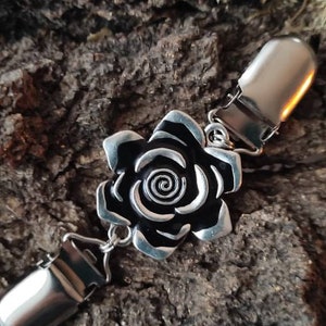 May include: A silver-coloured metal brooch featuring a black rose design. The rose has detailed petals and a spiral centre. The brooch has two metal clips on either side, connected by small chains. The background is a textured, brown surface.